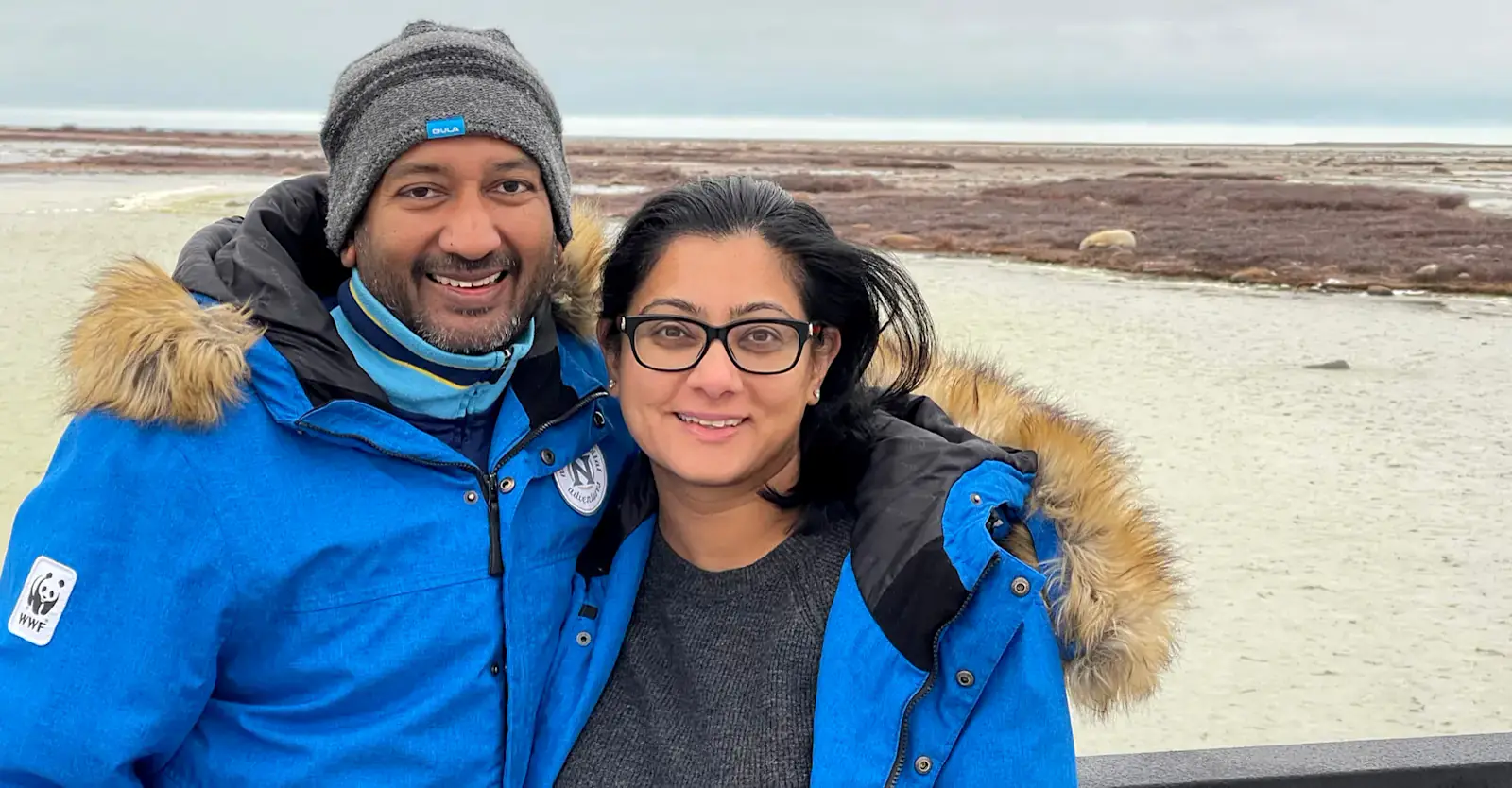 Two travelers in parkas with a polar bear on the tundra behind them, Churchill, Manitoba